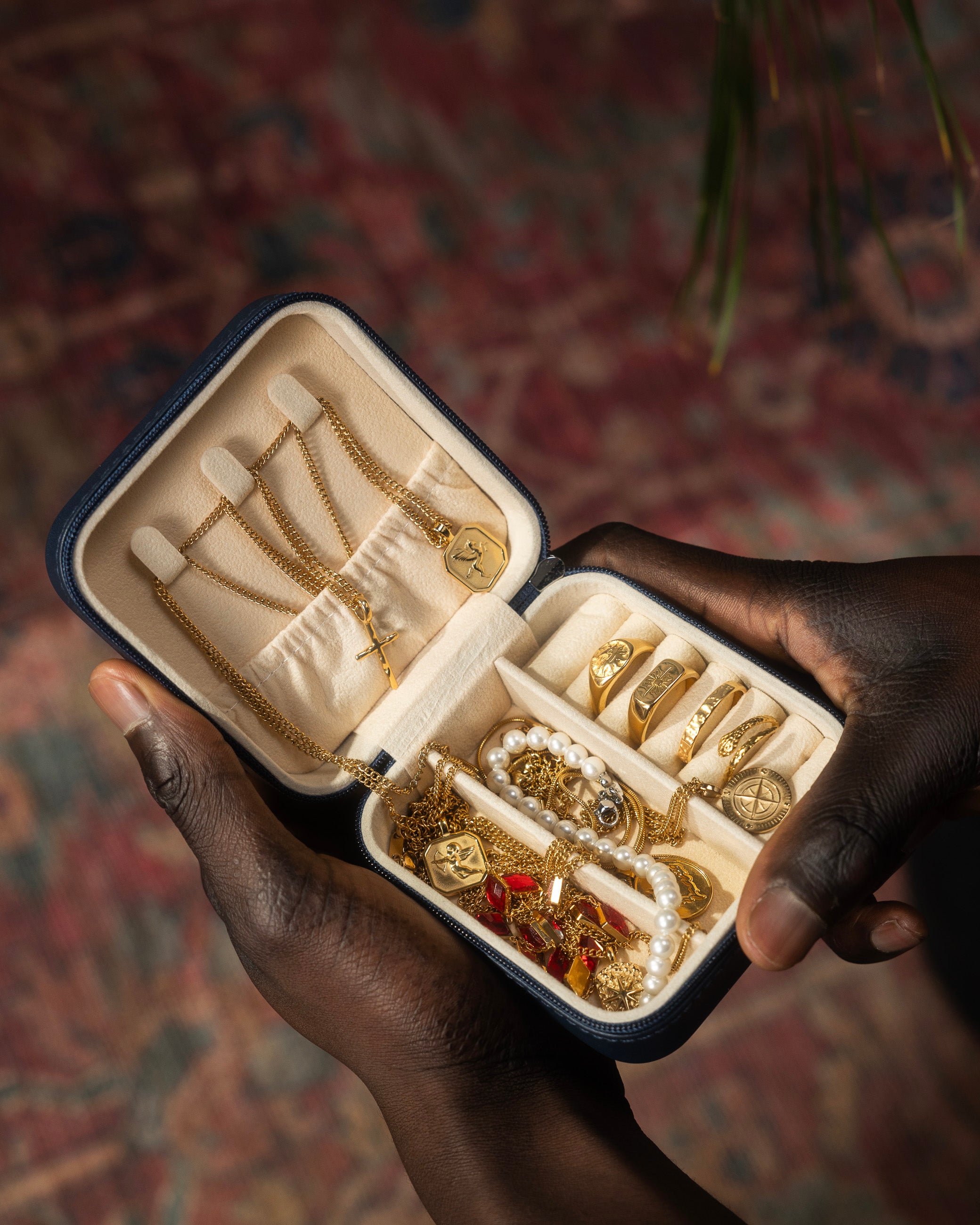Hands open a travel jewelry case displaying silver chains and a pearl bracelet on a textured grey surface.
