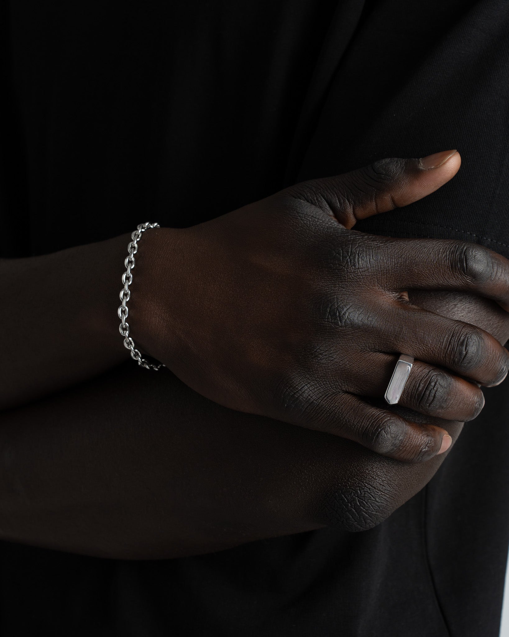 Wrist with a silver anchor bracelet and a rectangular ring on the finger against a dark outfit