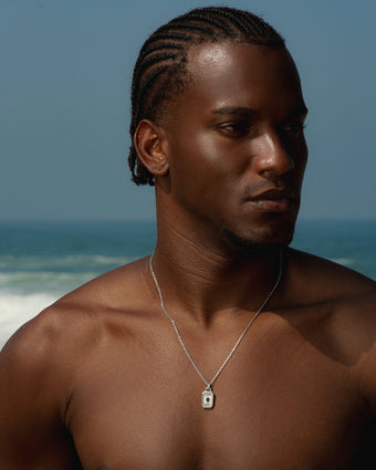 Man wearing a silver AETHER necklace with a square pendant against a seaside backdrop.