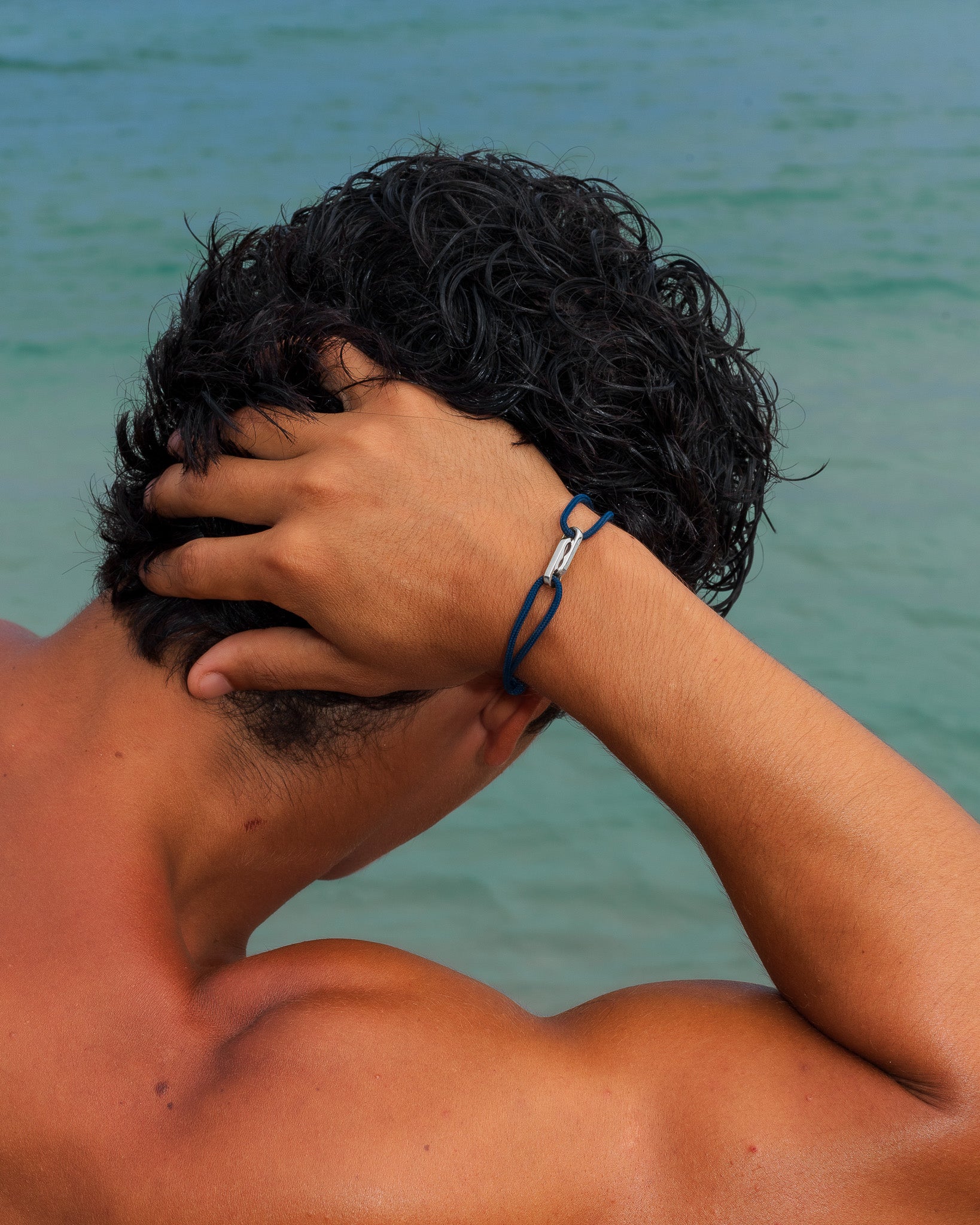 Vista de espaldas de un hombre sin camiseta con una pulsera de cordón azul y cierre de plata en la muñeca, con el mar de fondo.