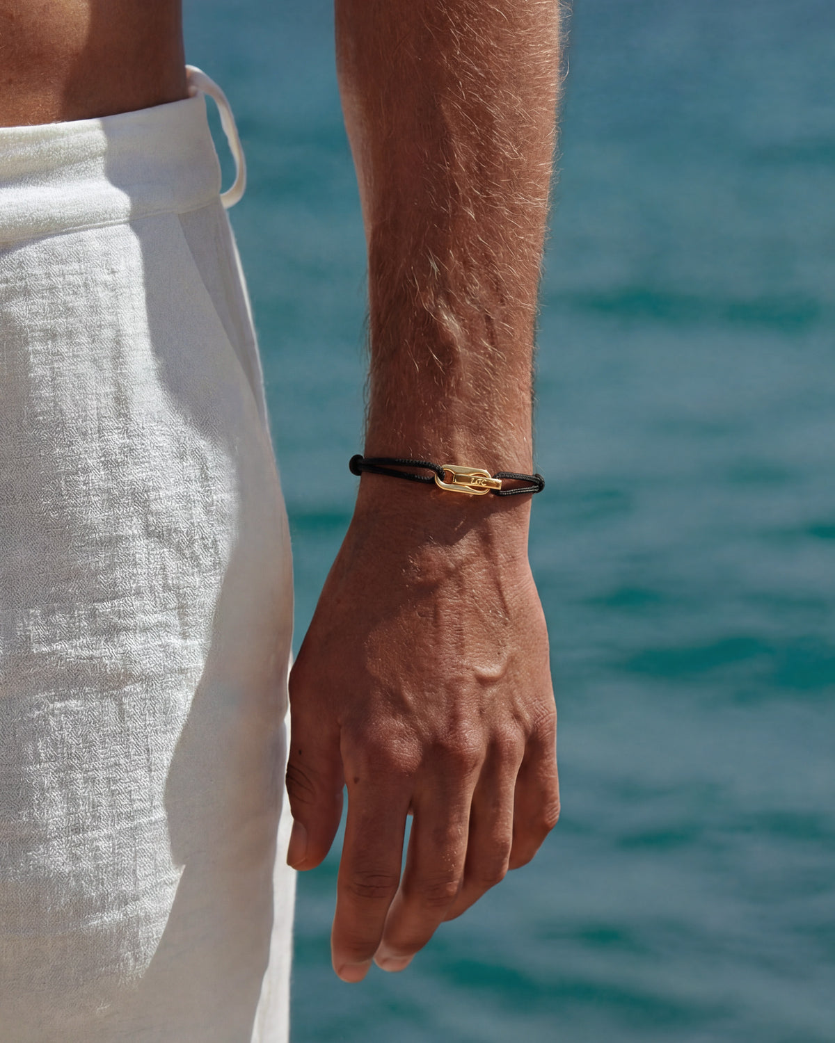Pulsera dorada Atlantis con cordón negro y cierre dorado en la muñeca junto al mar