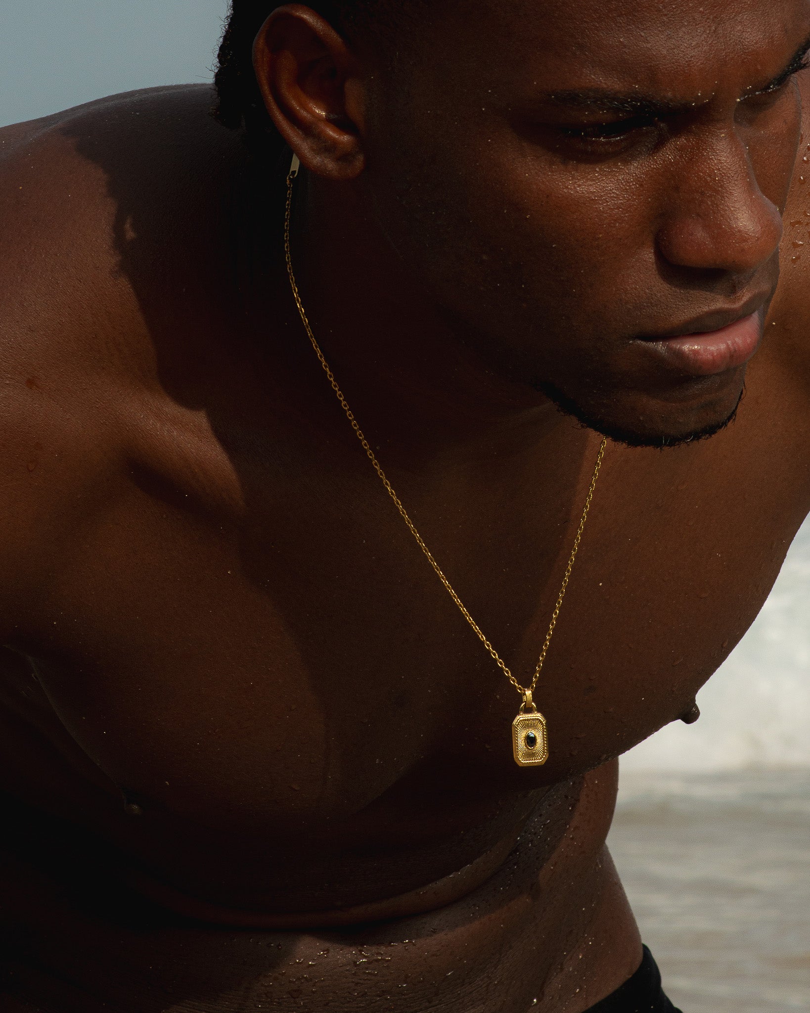 Close up of a person wearing a gold chain necklace with a rectangular pendant featuring a central dark stone