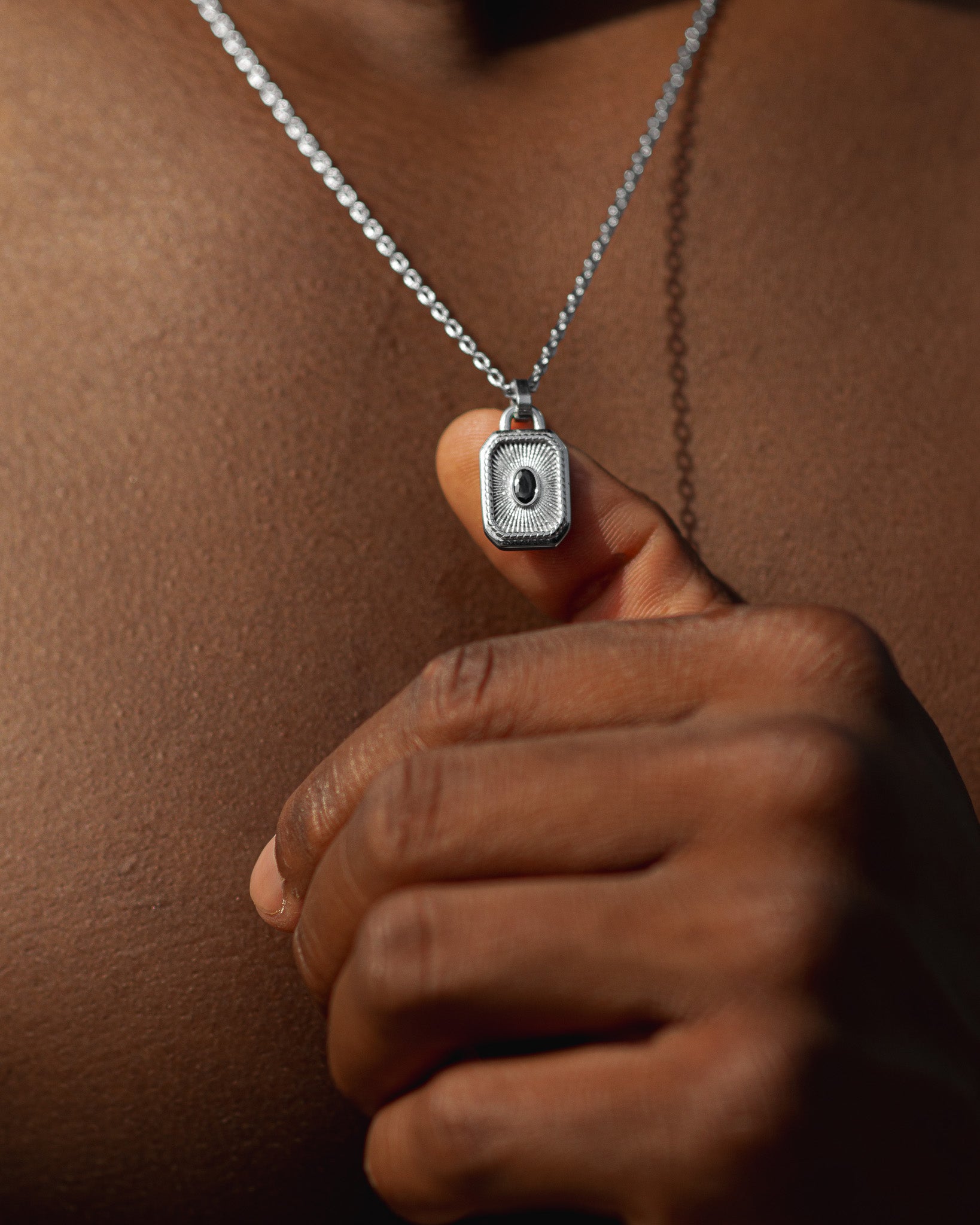 Man wearing a silver chain necklace with a rectangular pendant featuring a dark center near the ocean.
