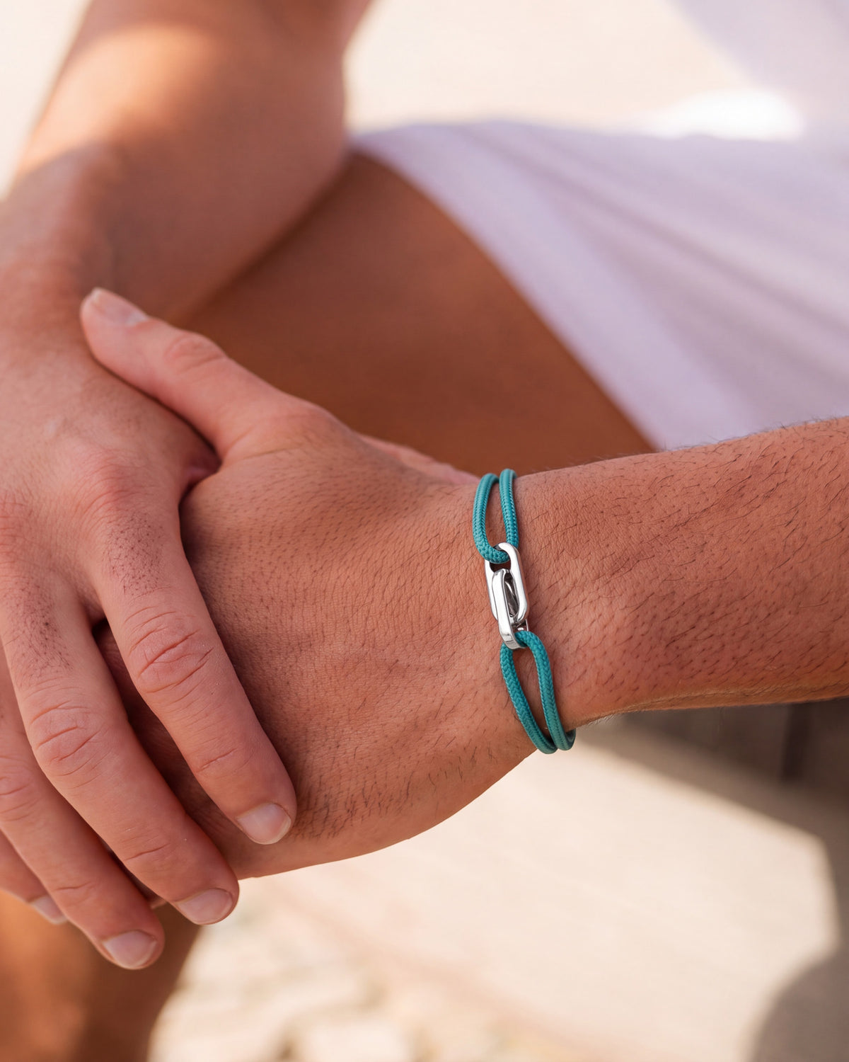 Atlantis Silberarmband mit blaugrüner Kordel am Handgelenk, die Hände ruhen im Sonnenlicht ineinander
