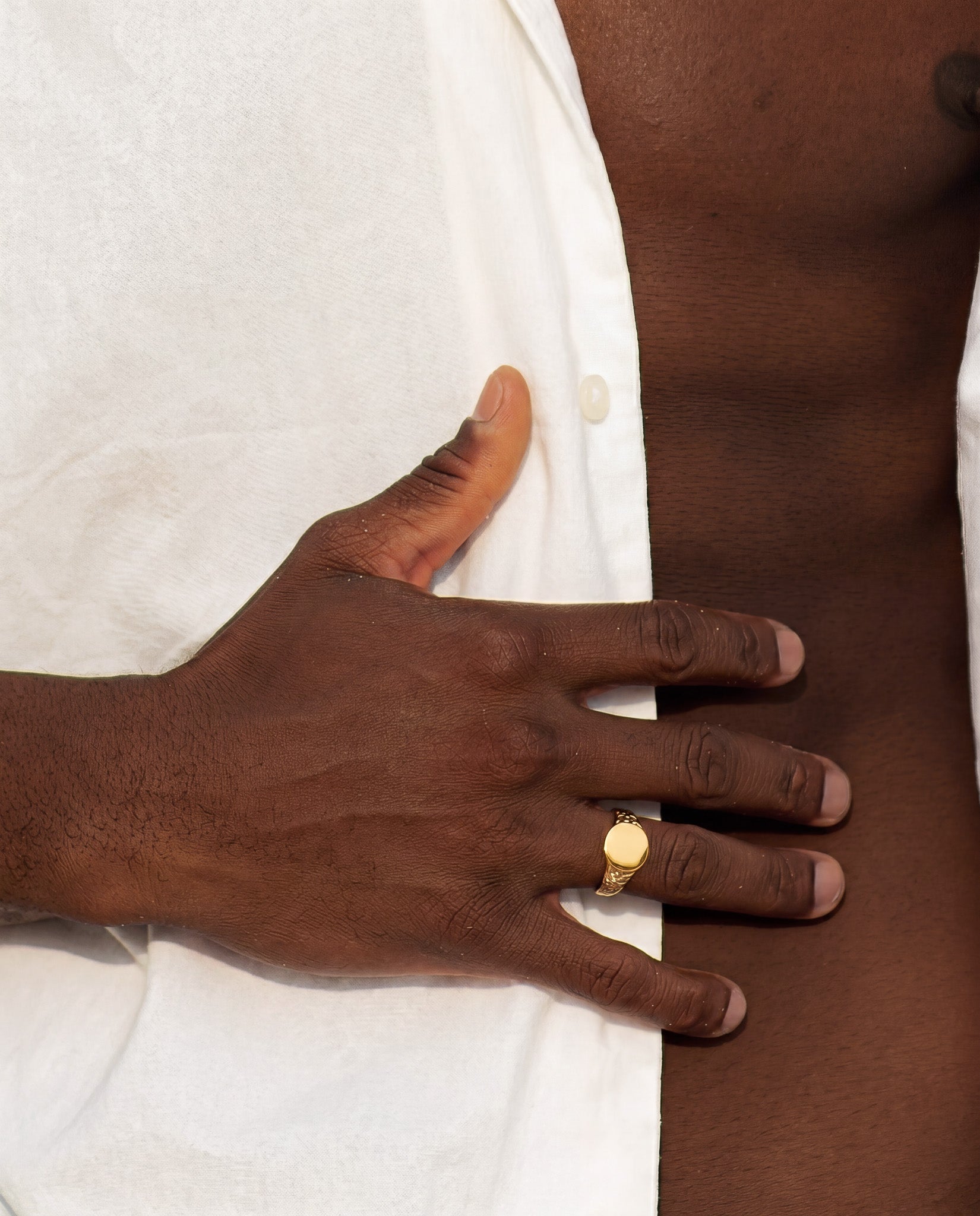 Gold Eden Ring with a smooth circular face on the ring finger of a dark-skinned hand resting on white fabric.