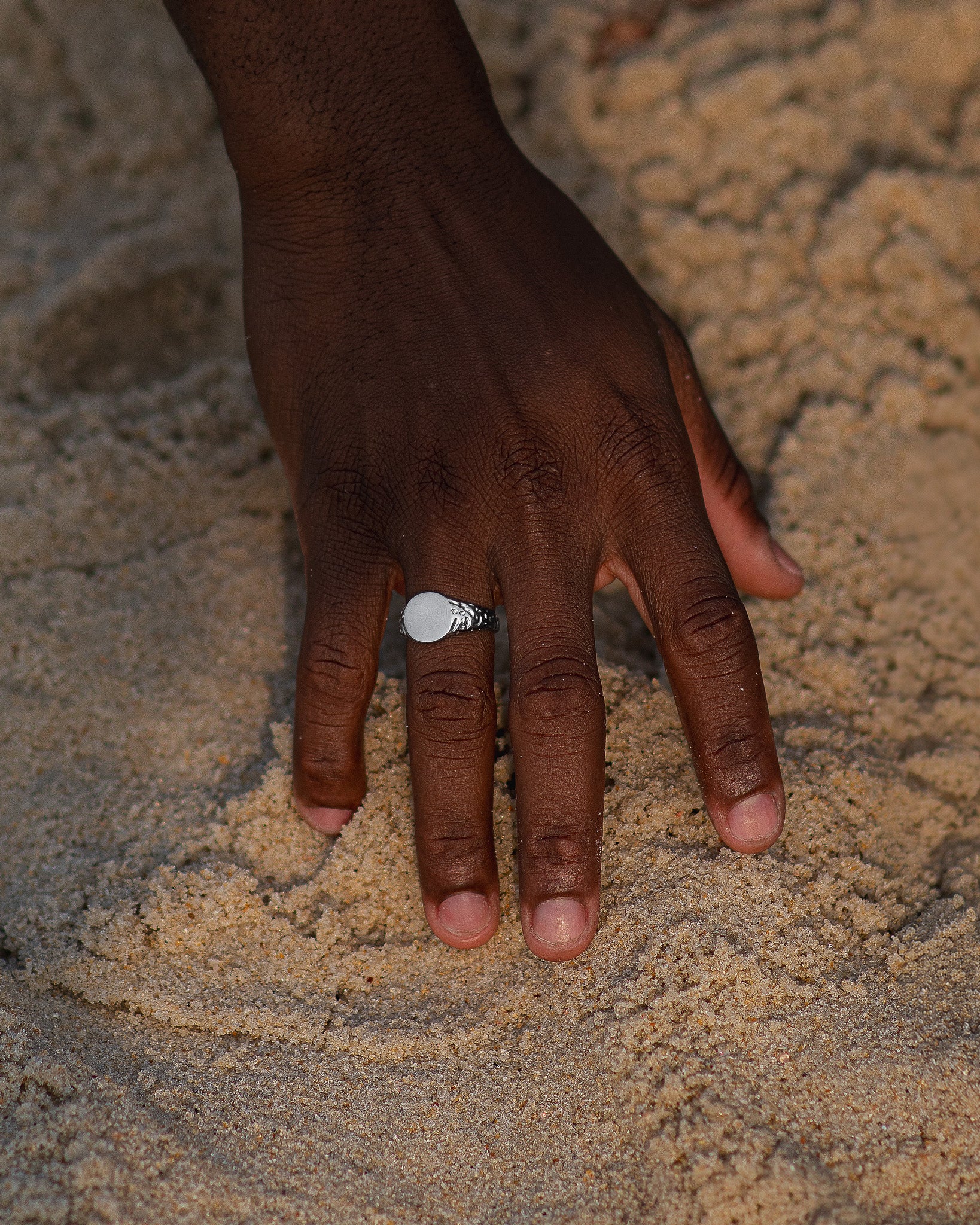 Front view of a silver EDEN ring with a smooth circular face on the wearer's finger against a sandy background.