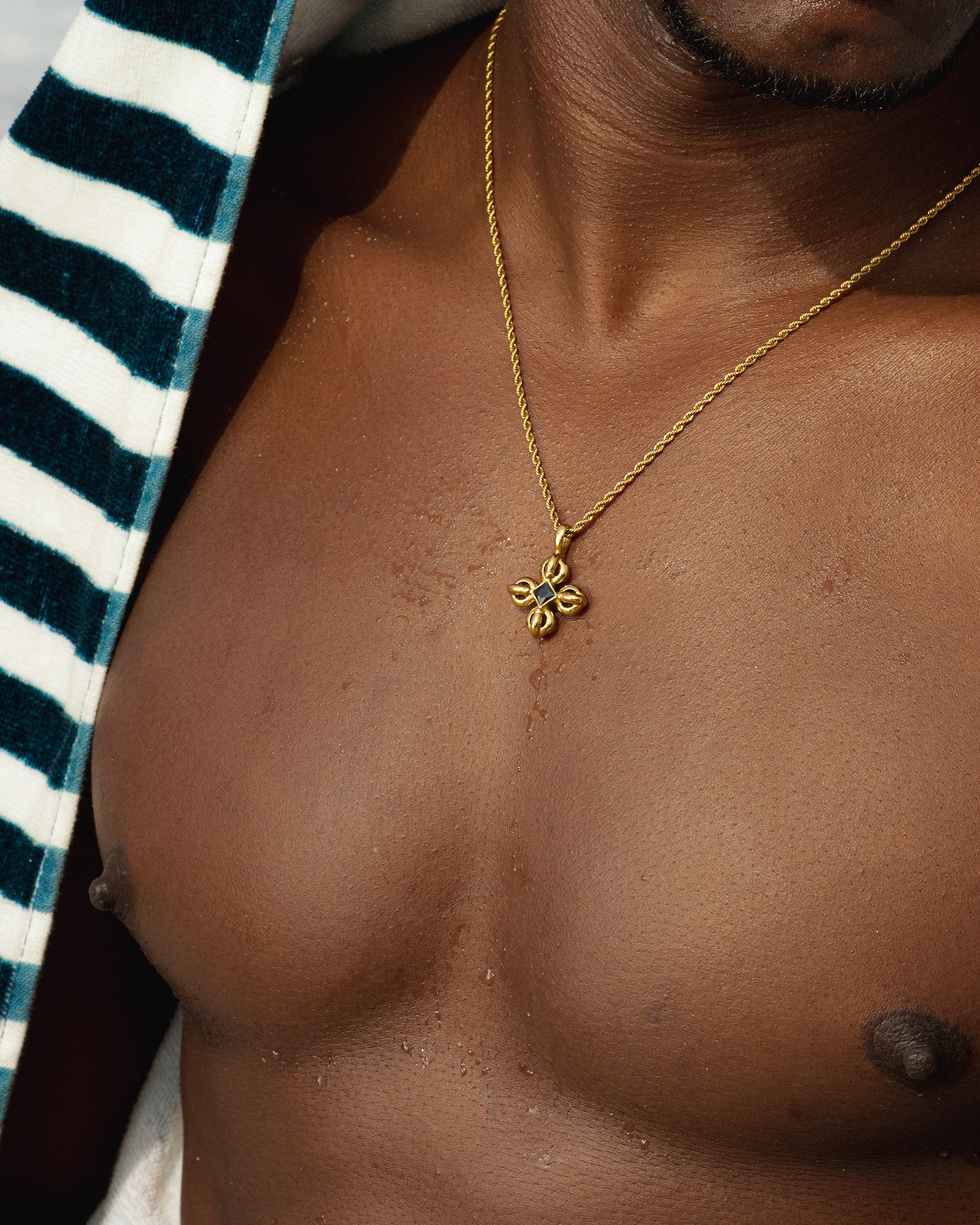 Gold flare necklace resting on a model's bare chest beside a striped blue and white garment.