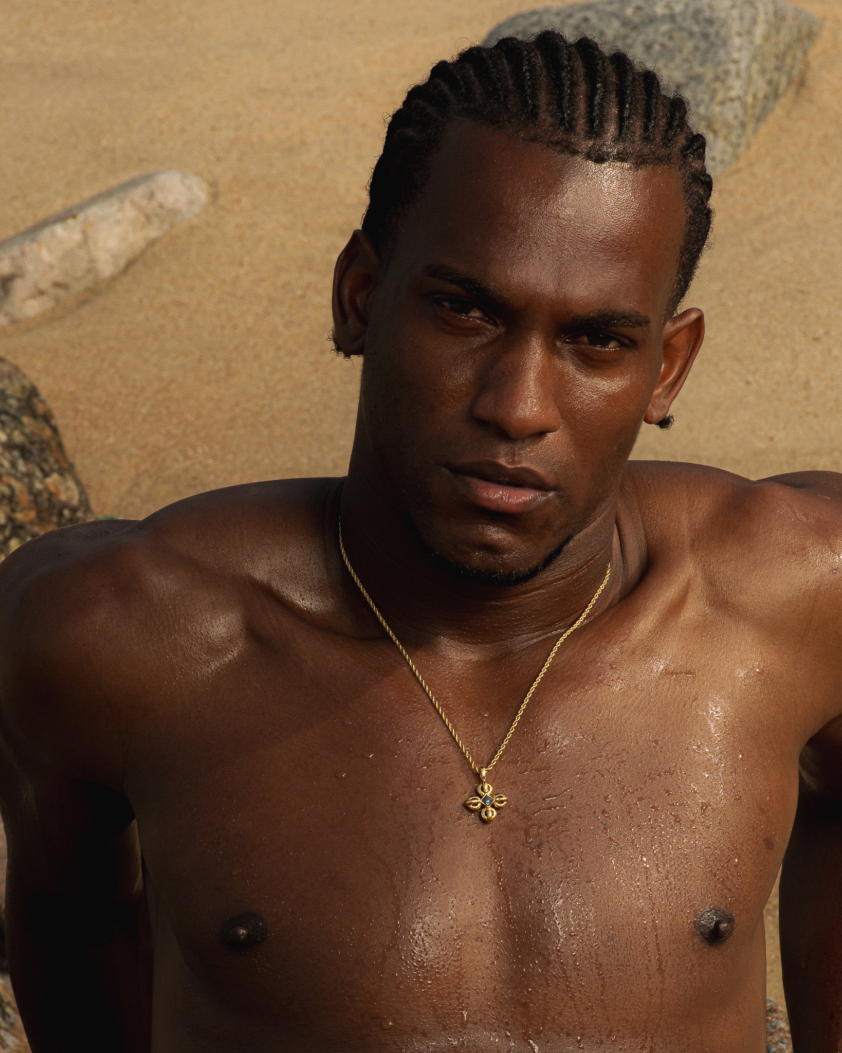 Portrait of a man with braided hair wearing a gold necklace against a sandy outdoor background.