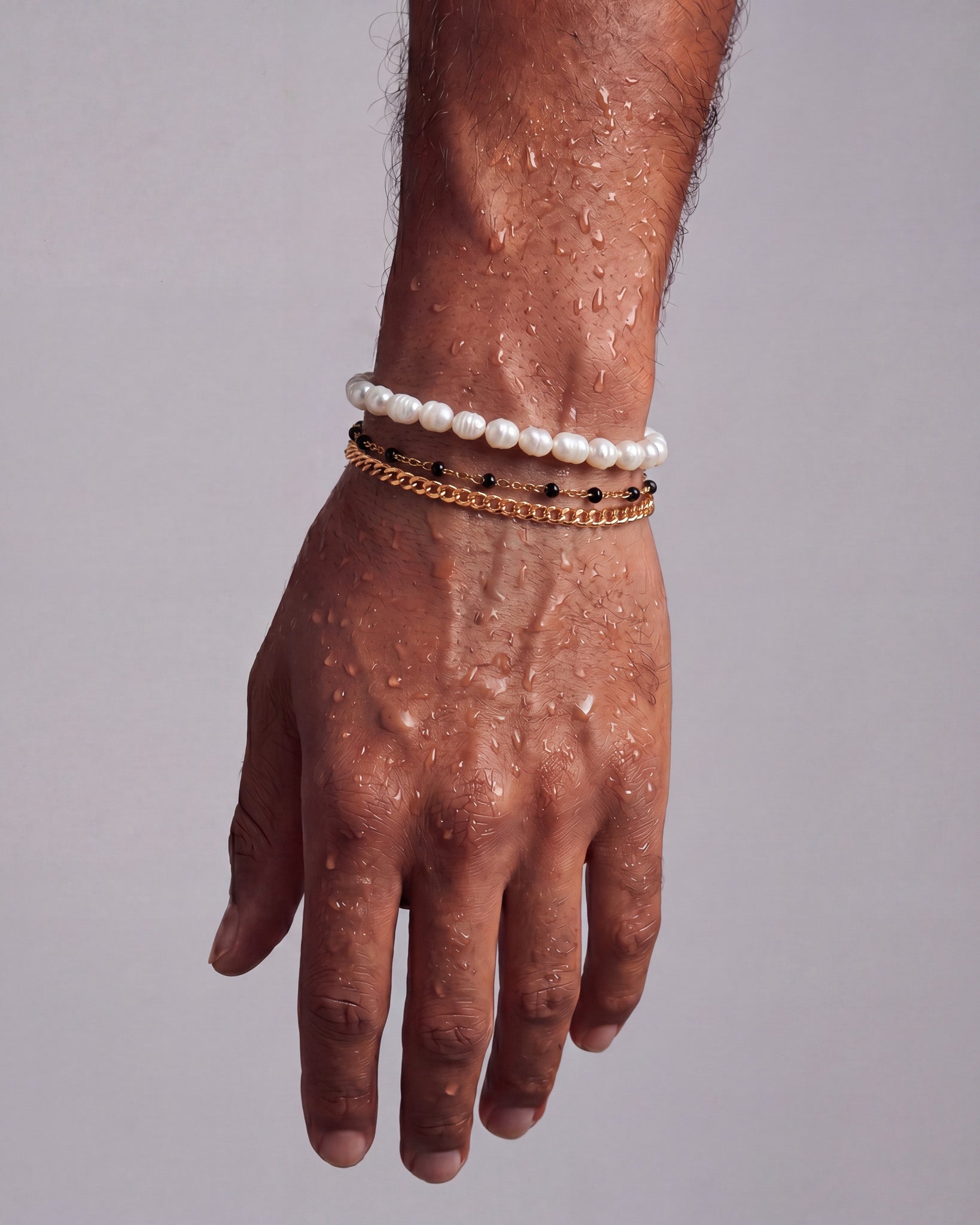 Wrist stacked with a white pearl bracelet and multiple gold chain bracelets against a pale backdrop.