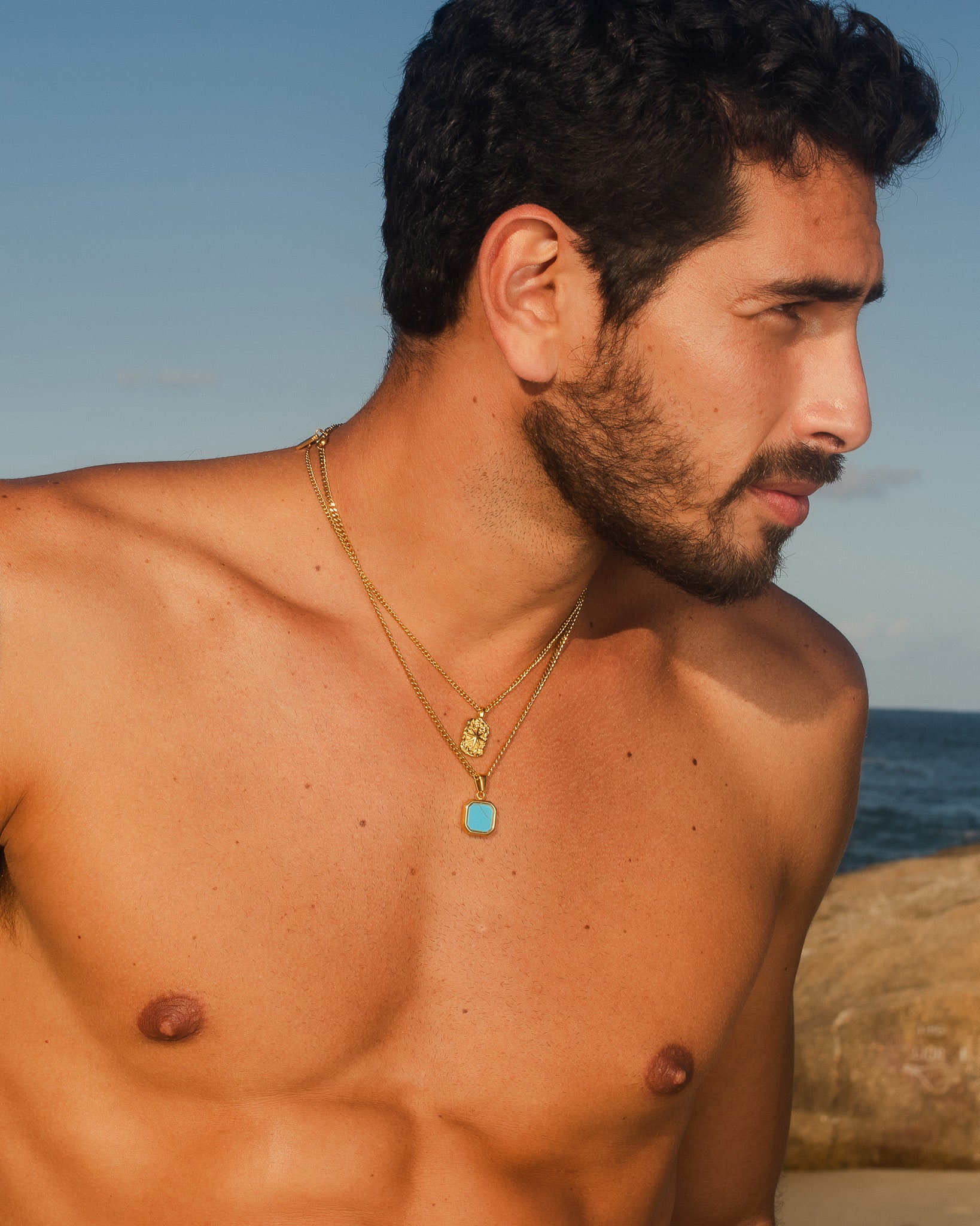 Close-up of a man wearing layered Nova Gold necklaces with a turquoise rectangular pendant and a small gold charm