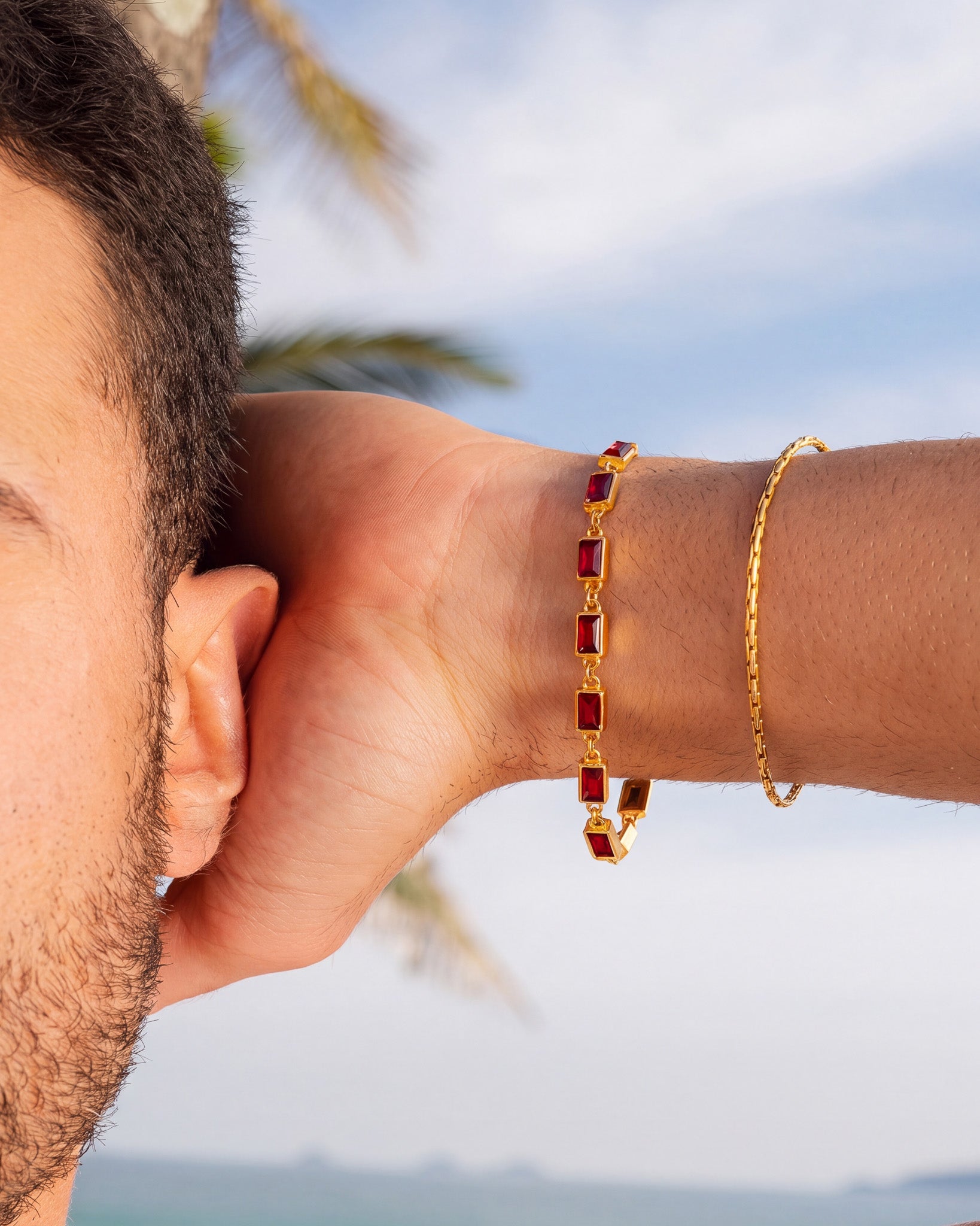 OMEN Gold Bracelet with red rectangular gemstones worn on a wrist against a blue sky