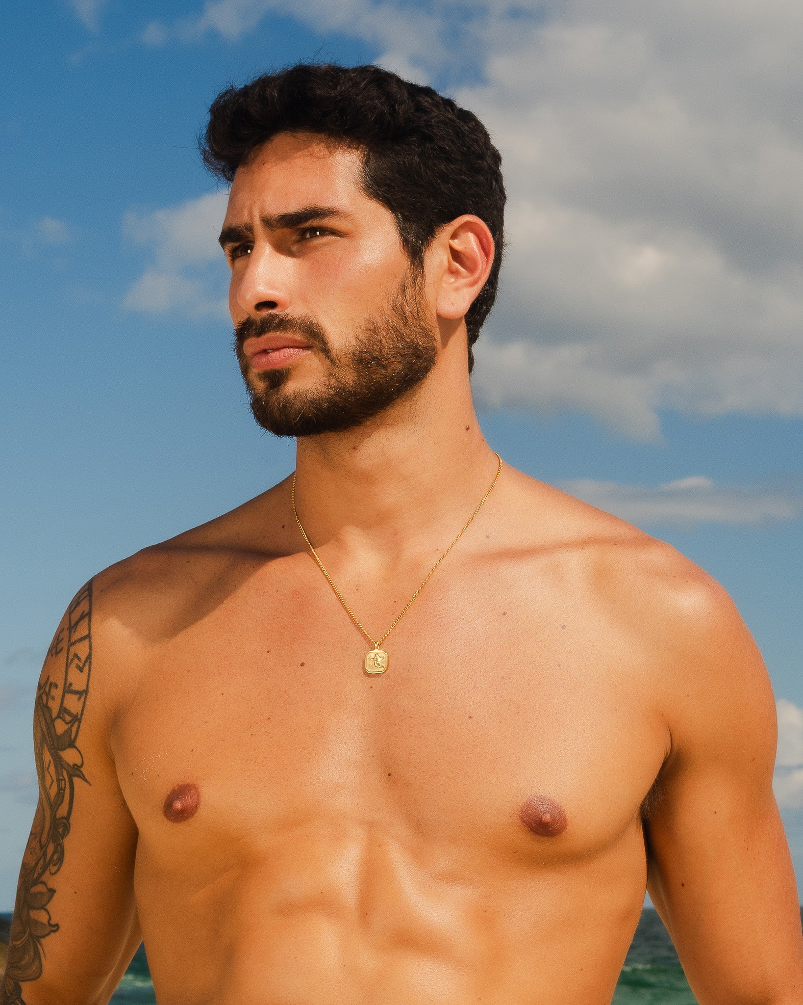 Man wearing a Savior gold necklace with a square pendant against a blue sky.